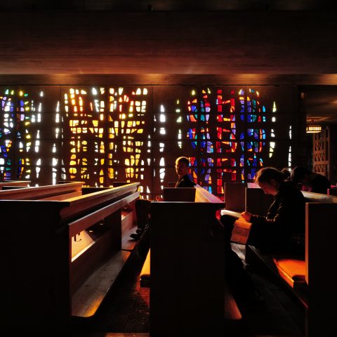 People sit quietly on wooden pews in a dimly lit church, with colorful light shining through stained glass windows behind them.