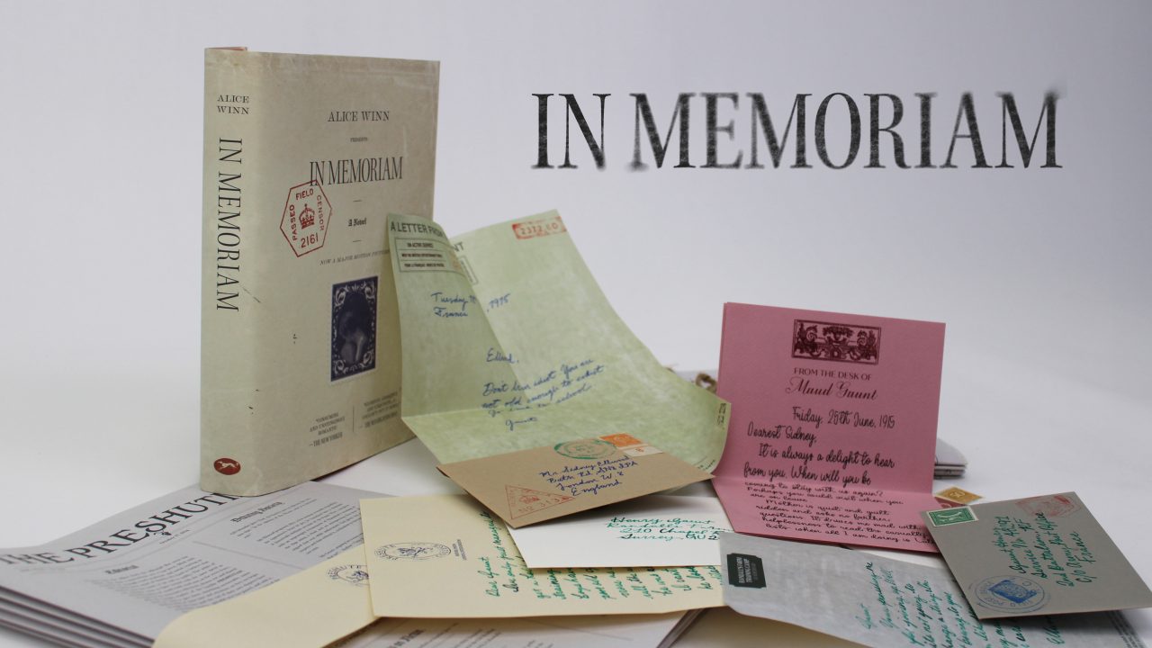 A book titled In Memoriam stands upright beside several open envelopes, handwritten letters, and printed documents arranged on a white surface. The words IN MEMORIAM appear in large text.