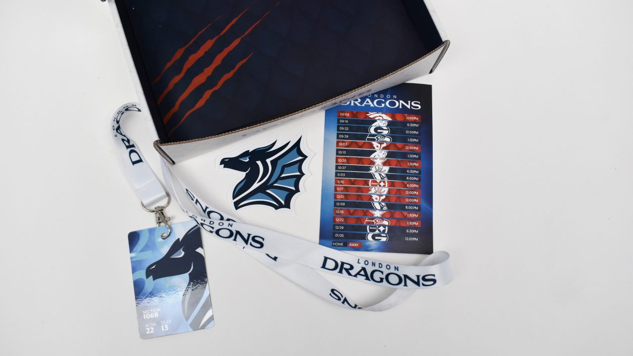 A box with a dragon logo, a London Dragons lanyard with an ID badge, and a schedule card featuring dates and times for various events displayed on a white surface.