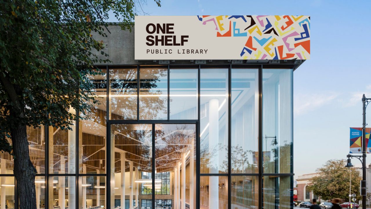 Glass-walled modern building with a sign reading One Shelf Public Library above the entrance; street and trees visible outside.