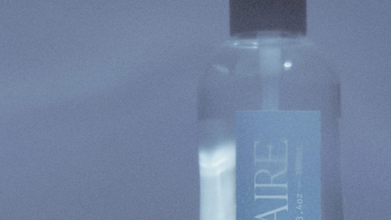 A clear spray bottle labeled SOLAIRE Room Spray 3.4oz with a black cap sits against a light blue background.