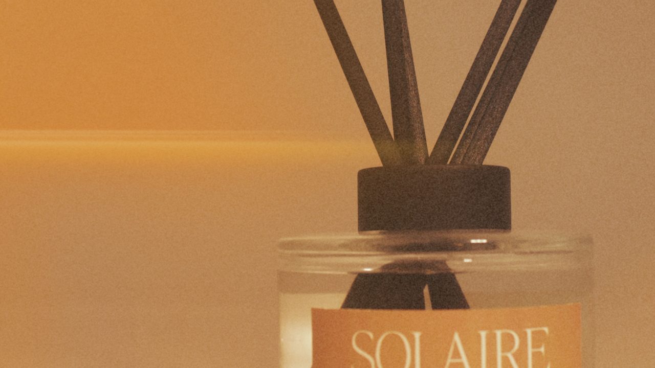 A glass reed diffuser labeled SOLAIRE with several black reeds sticks out, set against a warm, softly lit background.