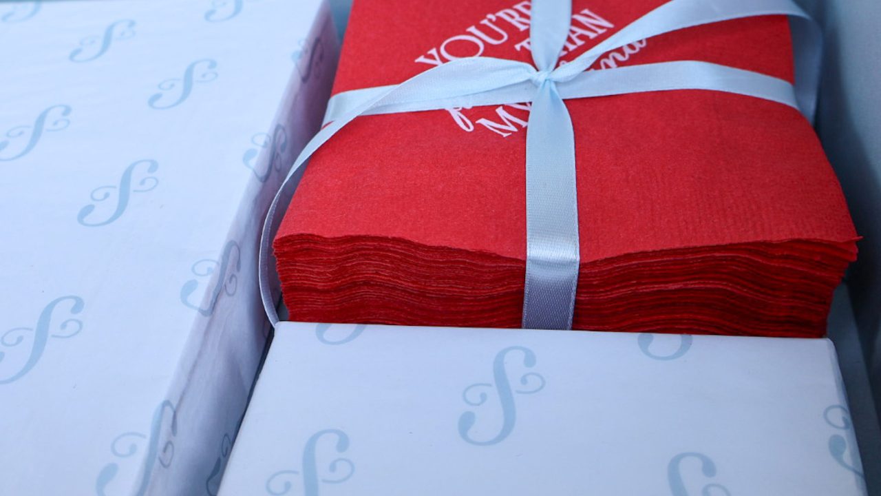 A stack of red napkins tied with a light blue ribbon next to white boxes printed with a light blue cursive letter S inside a box.