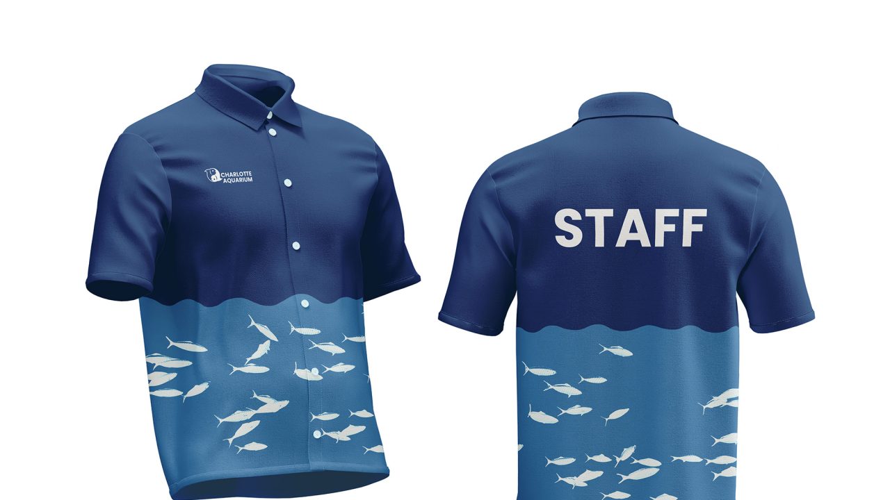 Two blue staff shirts with ocean and fish designs; one shows the front with a logo, the other shows the back with STAFF printed in white uppercase letters.