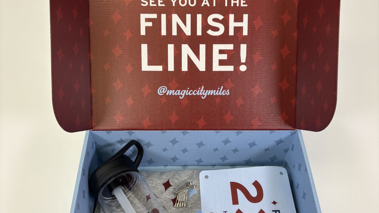 Open box with a water bottle, a gray T-shirt, a half marathon race bib labeled RICHARD 2307, and a medal; the lid reads See you at the finish line! @magiccitymiles.