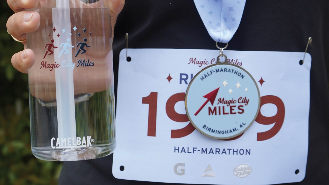 A person holds a CamelBak water bottle while wearing a race bib numbered 1989 and a Magic City Miles Half-Marathon finishers medal.