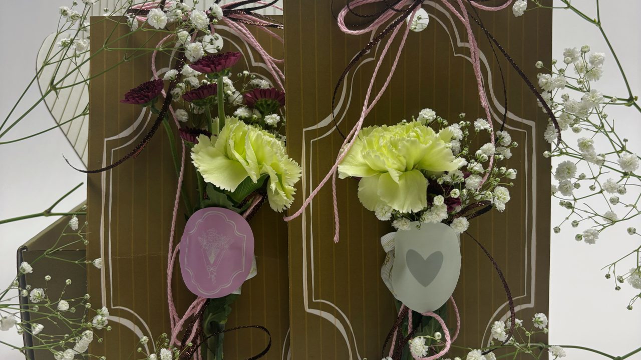 A decorative card stands upright, adorned with green carnations, baby’s breath, pink and black ribbons, and translucent plastic flower holders with heart and tulip motifs.