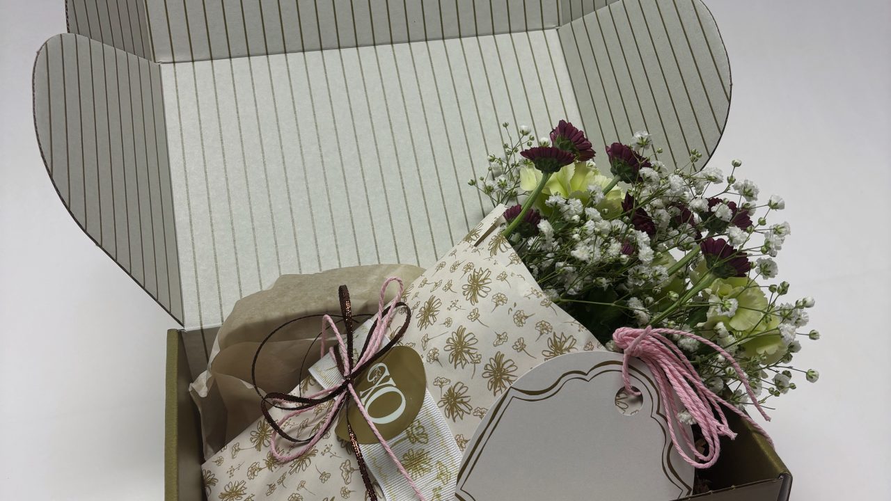 A brown gift box with an open lid contains a wrapped present, a blank tag with a string, and a small bouquet of purple and white flowers.