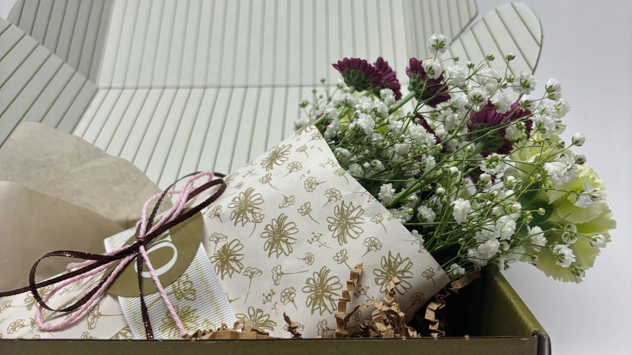 A small bouquet of flowers wrapped in patterned paper is placed inside an open olive green gift box with a logo on the front.