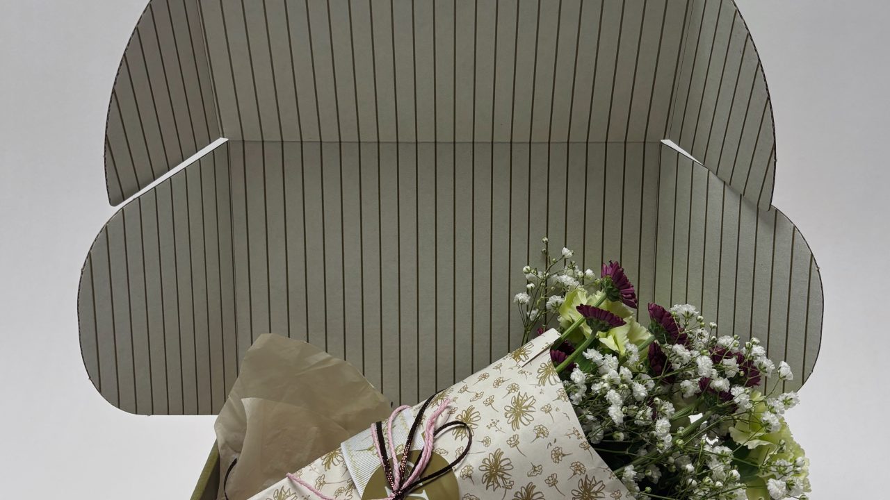 A small bouquet of flowers wrapped in patterned paper and tied with string sits inside an open, green-striped gift box.