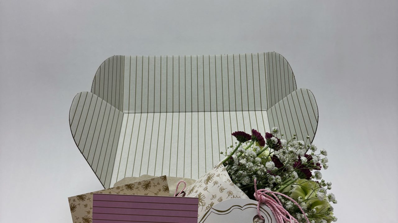 A gift box with striped interior contains a bouquet of flowers, a pink striped card, and a floral-designed envelope, all arranged neatly against a plain white background.