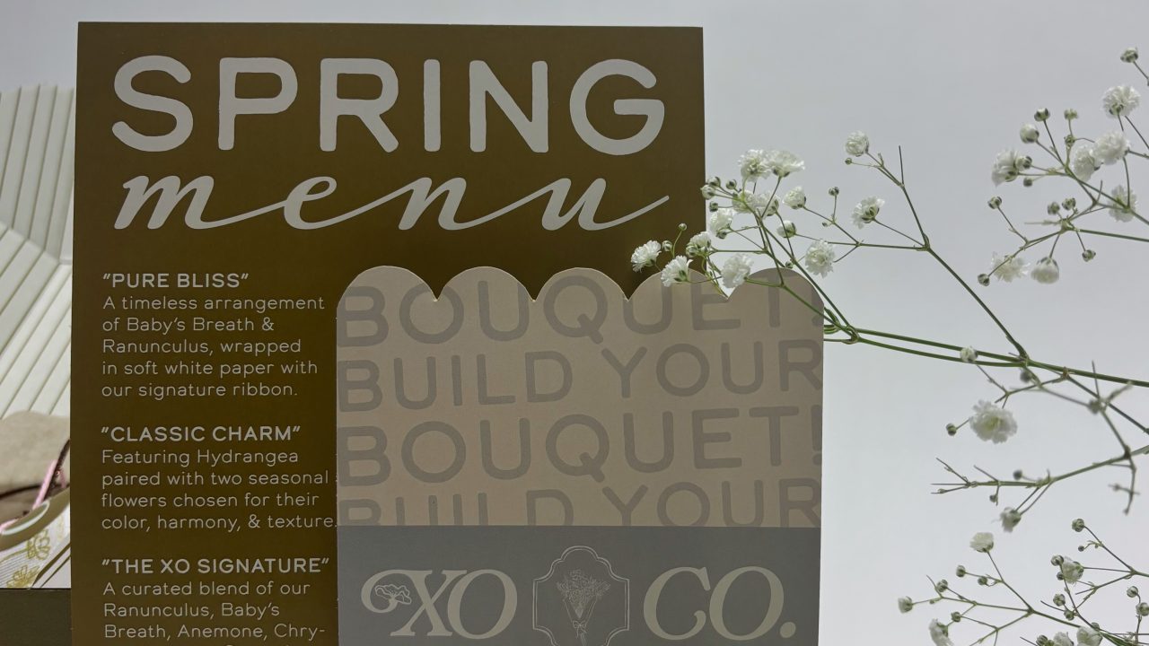 A spring menu card for XO, featuring flower arrangement options, is displayed upright with babys breath flowers beside it against a plain background.