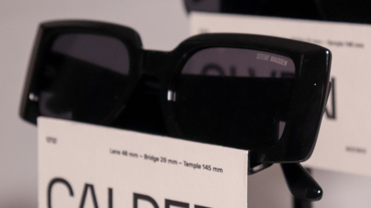 Two pairs of black sunglasses on display stands, each with a label; the front label reads CALDER and details lens, bridge, and temple measurements.