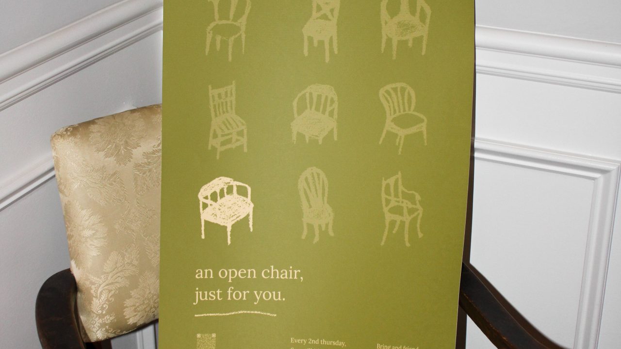 A green poster with illustrations of chairs rests on a beige upholstered chair, beneath a framed dolphin print on a white paneled wall.