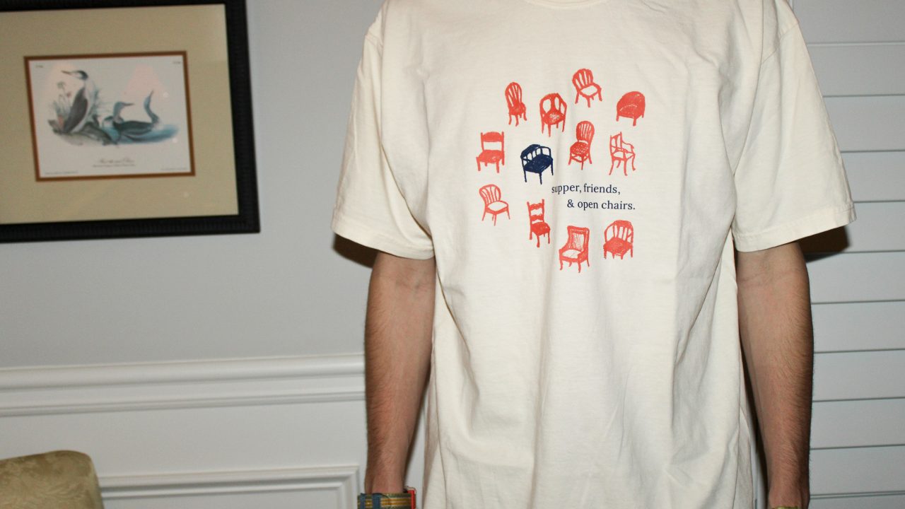 Person wearing a white t-shirt with illustrations of chairs and the text supper, friends, & open chairs printed on the front.