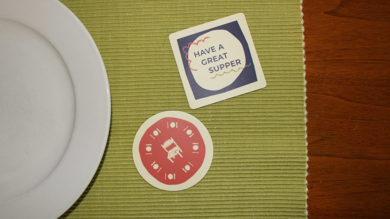 A white plate next to two coasters on a green placemat; one coaster says Have a great supper, and the other shows a table surrounded by utensils.