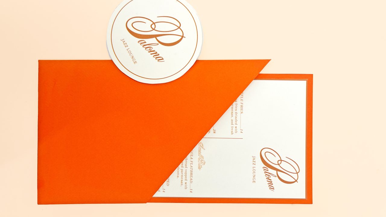 An orange envelope partially reveals a white card with gold writing and a matching round coaster, both labeled “Paloma” with stylized script.
