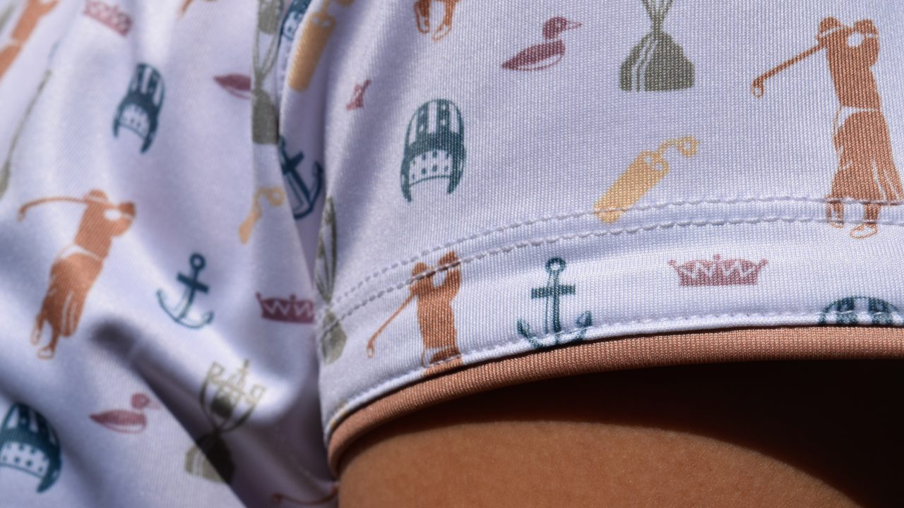 Close-up of a shirt sleeve featuring a pattern with images of golfers, golf clubs, trophies, anchors, and ducks in muted colors.
