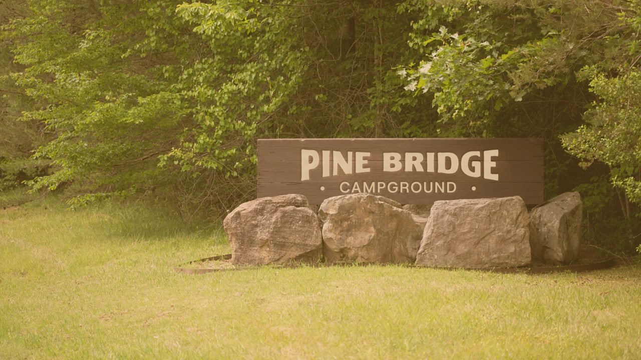 A wooden sign reading Pine Bridge Campground stands among large rocks and green foliage.