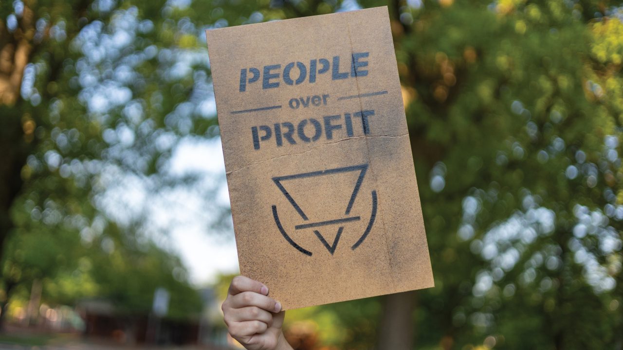 A hand holds a cardboard sign reading People over Profit with a stylized symbol, against a background of green trees.