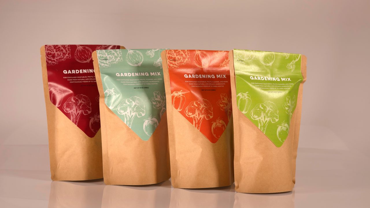 Four brown paper gardening mix bags stand side by side, each with a different colored label: red, green, orange, and yellow, featuring white vegetable illustrations.