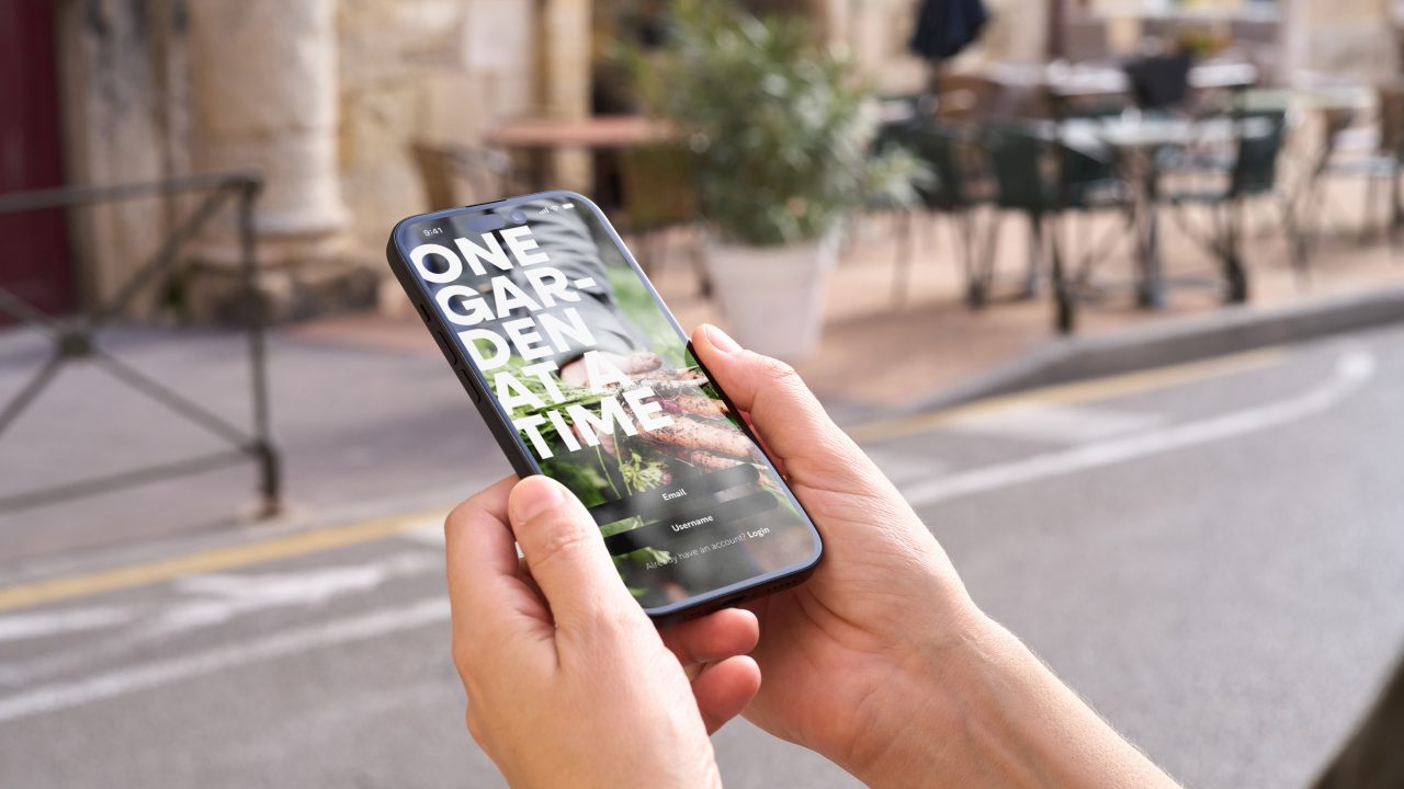 A person holds a smartphone displaying a website with the text ONE GARDEN AT A TIME on the screen, with an outdoor café in the blurred background.