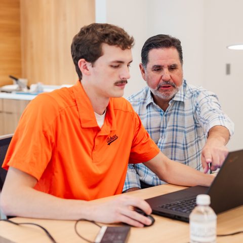 student working at laptop with professor, Alex Krumdieck, behind him instructing him