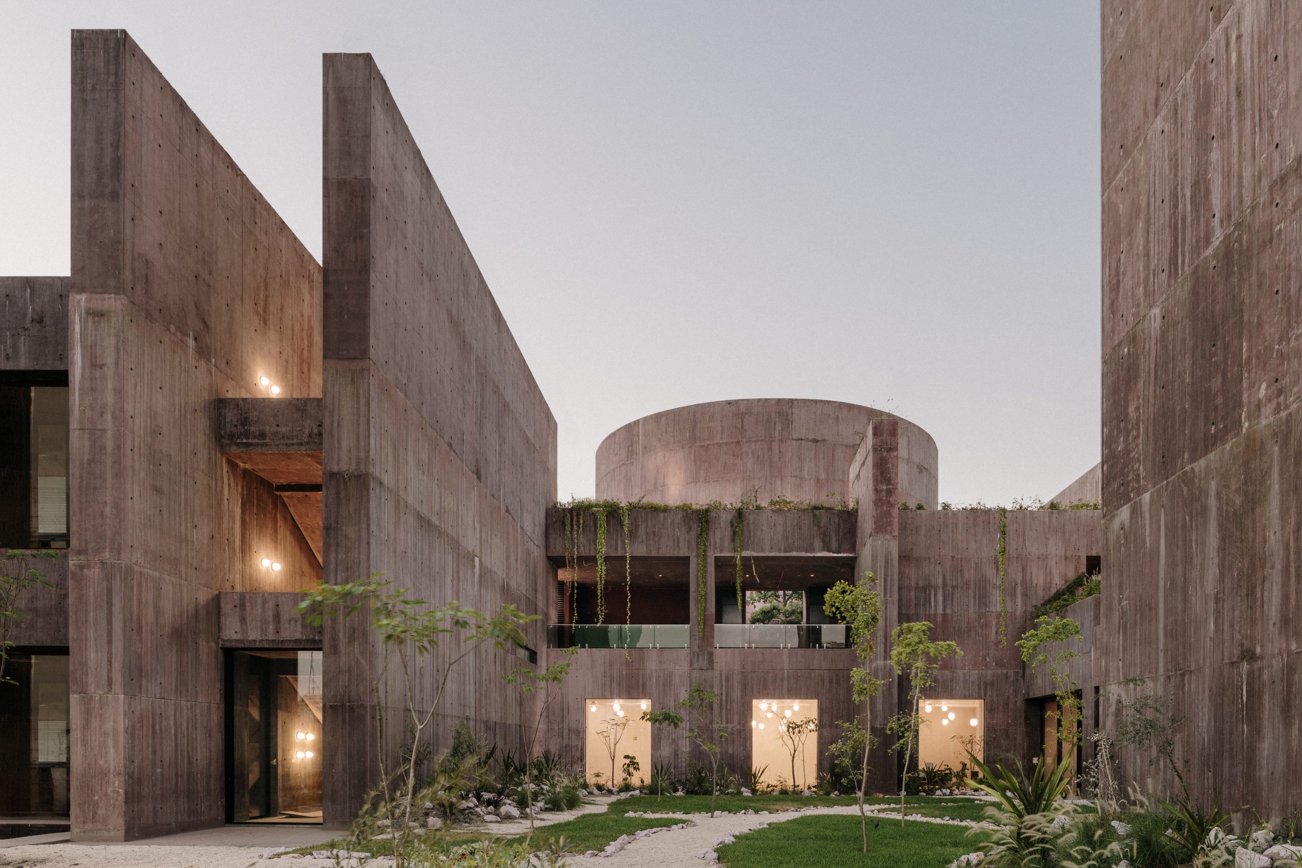 Concrete building with large, angular walls and a cylindrical structure. Soft lighting visible through windows, surrounded by trees and plants.