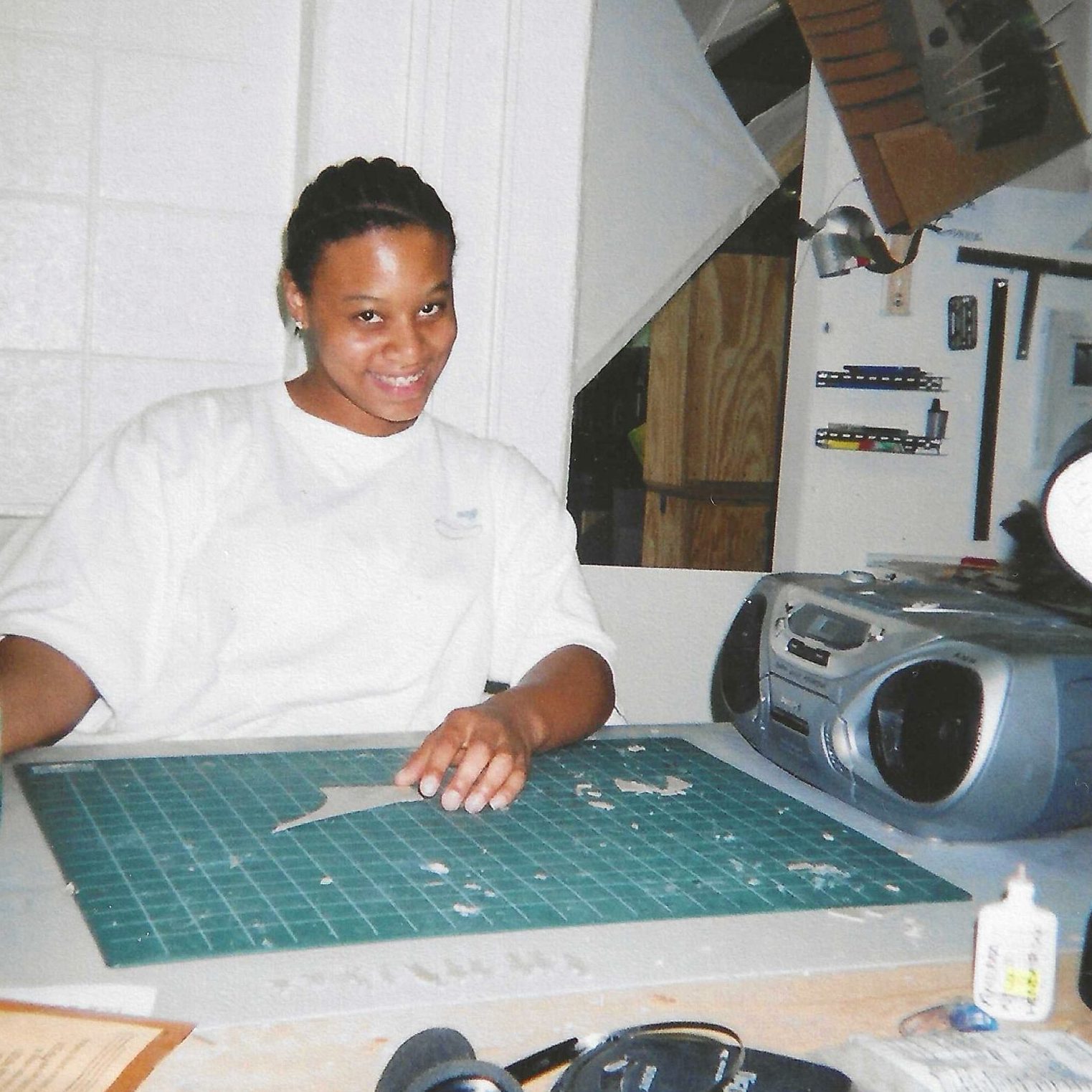 A person sits at a worktable with a green cutting mat, using a craft knife. A stereo, glue bottle, and various tools are on the table. The person is smiling at the camera.