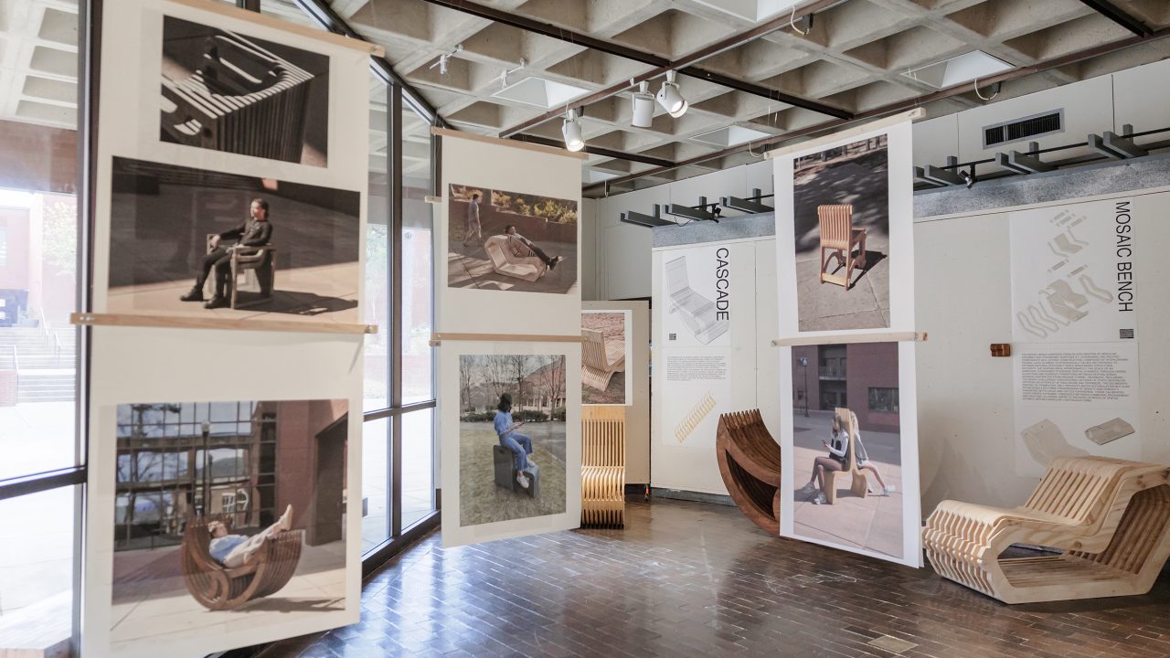 Auburn University Environmental Design student work Art exhibit room with large photos of people sitting on modern wooden benches and chairs, displayed alongside the actual furniture pieces and design descriptions on the walls.