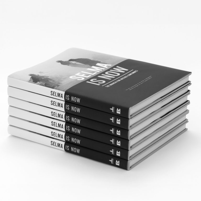 A stack of six hardcover books titled Selma Is Now with a black-and-white cover image of people marching, neatly arranged on a white surface.