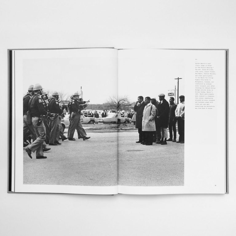 Black-and-white photo in a book showing police officers facing a group of civilians standing in a line; cars and trees are visible in the background.