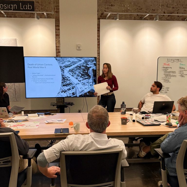 A woman stands and presents a slide titled “Death of Urban Centers Post World War II” to four seated colleagues in a modern meeting room.