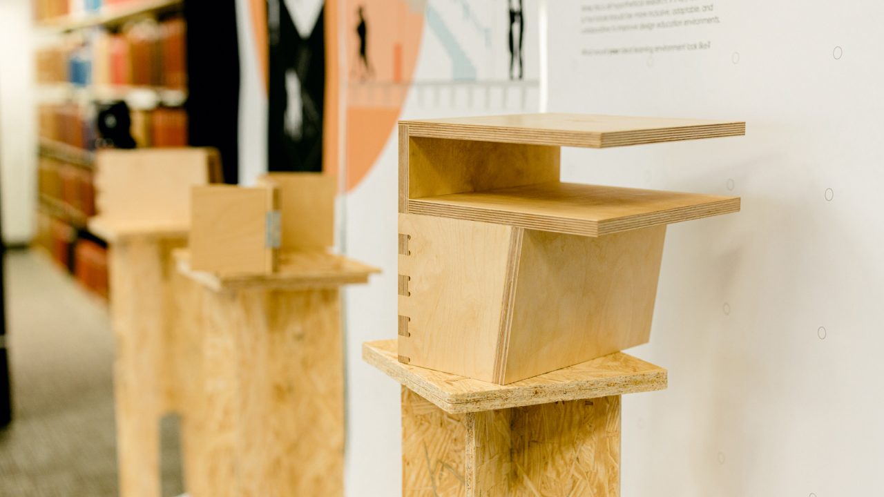 Auburn University Environmental Design student work A modern wooden architectural model with layered sections is displayed on a plywood pedestal, with similar models visible in the background.