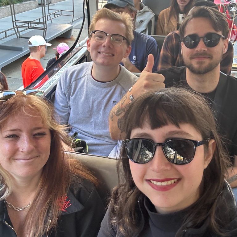 Four people sit closely together on an outdoor bus or tram, smiling at the camera. One person gives a thumbs-up. Other passengers are visible in the background.