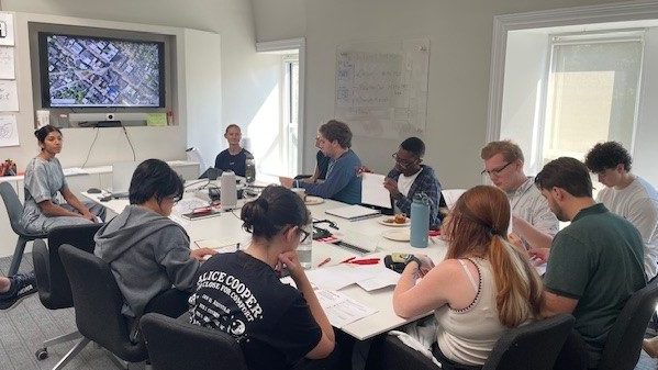 A group of people sit around a conference table in a bright room, looking at documents and a screen displaying an aerial map.