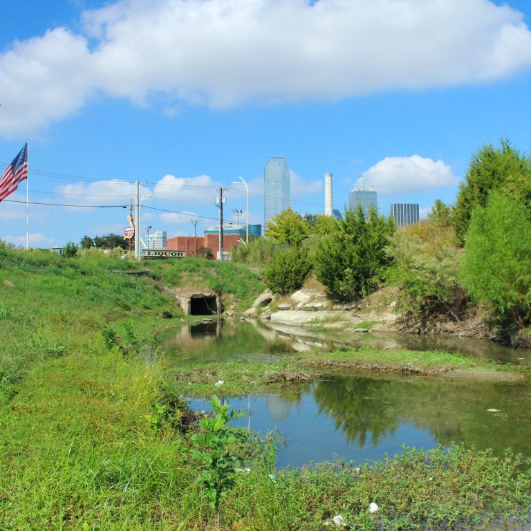 A small creek with overgrown banks flows toward a tunnel, with city buildings and an American flag visible in the background under a blue, cloudy sky.