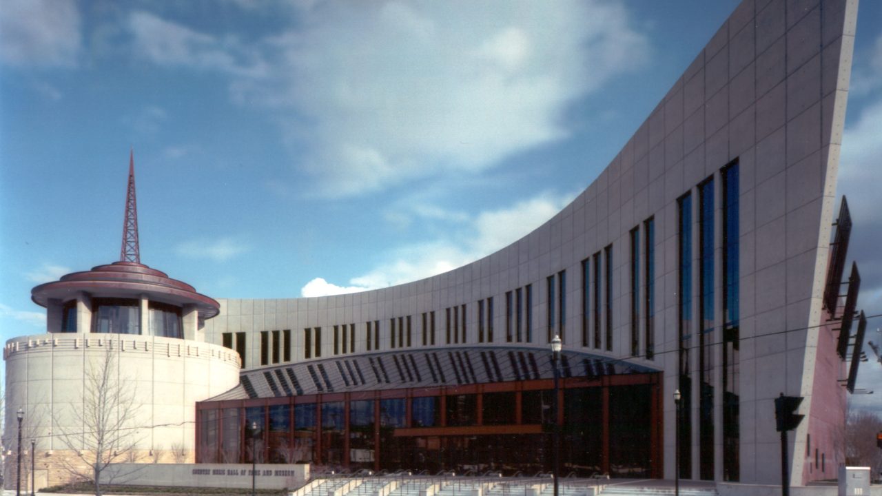 Modern building with a curved glass facade and a tower with a spire, set against a partly cloudy sky.