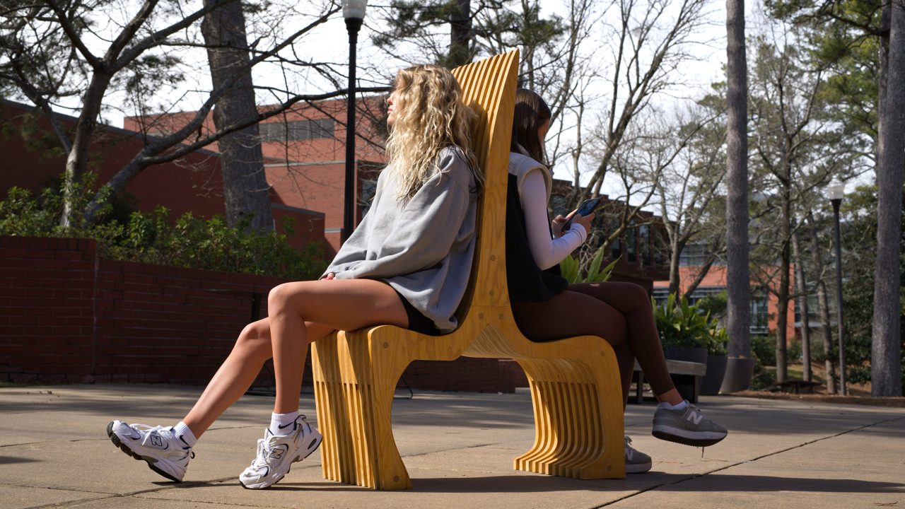 Auburn University Environmental Design student work Two people sit back-to-back on a uniquely shaped wooden bench outdoors, both wearing athletic clothing and sneakers, with trees and a brick building in the background.