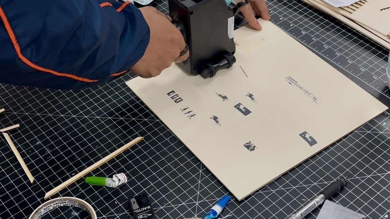 Auburn University Environmental Design student work Person operating a handheld printer to print text on a sheet, surrounded by various tools and materials on a cutting mat.