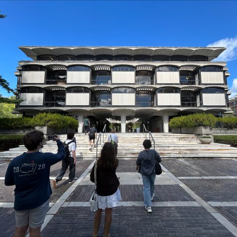 Several people walk toward a modern, multi-story concrete and glass building with cantilevered upper floors on a sunny day.