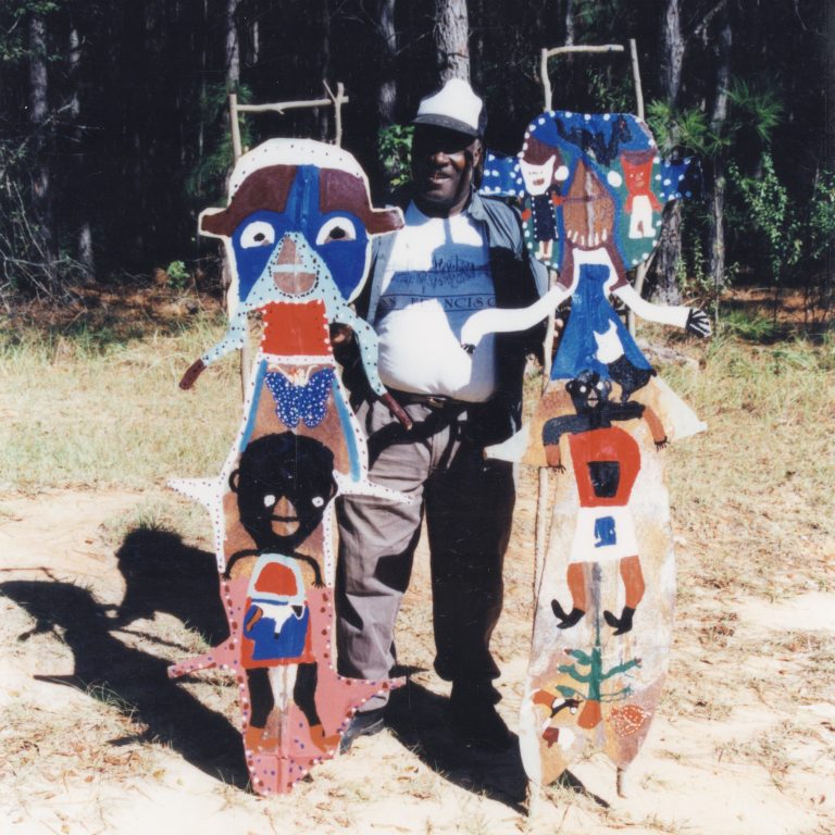 A man stands outdoors holding two tall, colorful, abstract wooden sculptures featuring faces and patterns, with trees in the background.