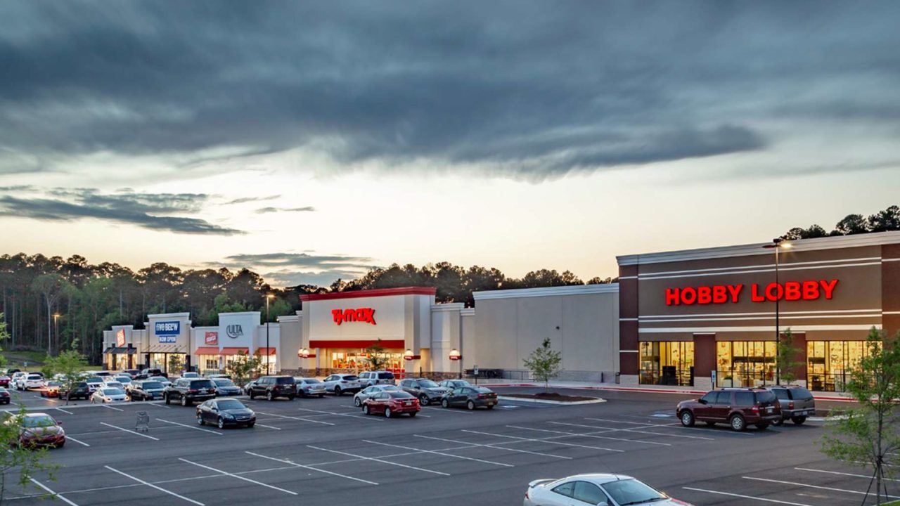 A shopping center parking lot with several cars and storefronts, including Hobby Lobby, TJ Maxx, and other retailers, at dusk.