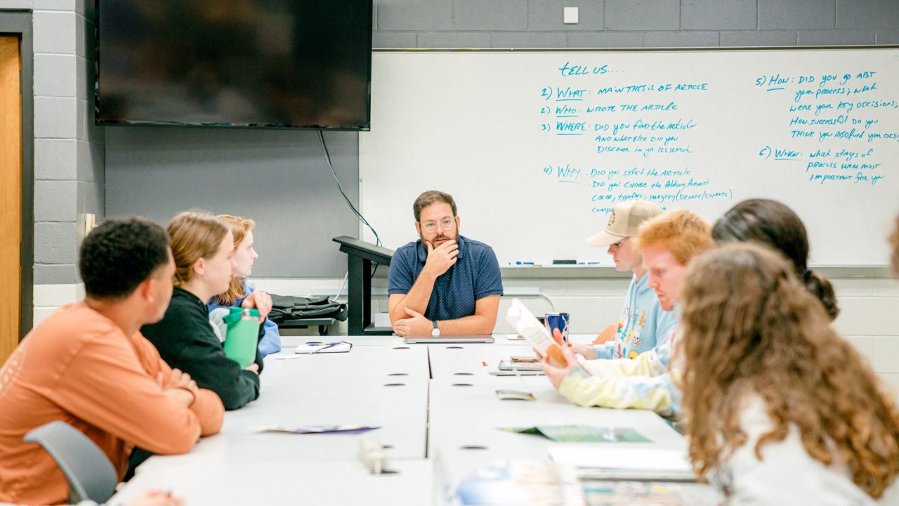 A group of students sit around a table listening to a man speaking at the front of a classroom with a whiteboard and TV on the wall.