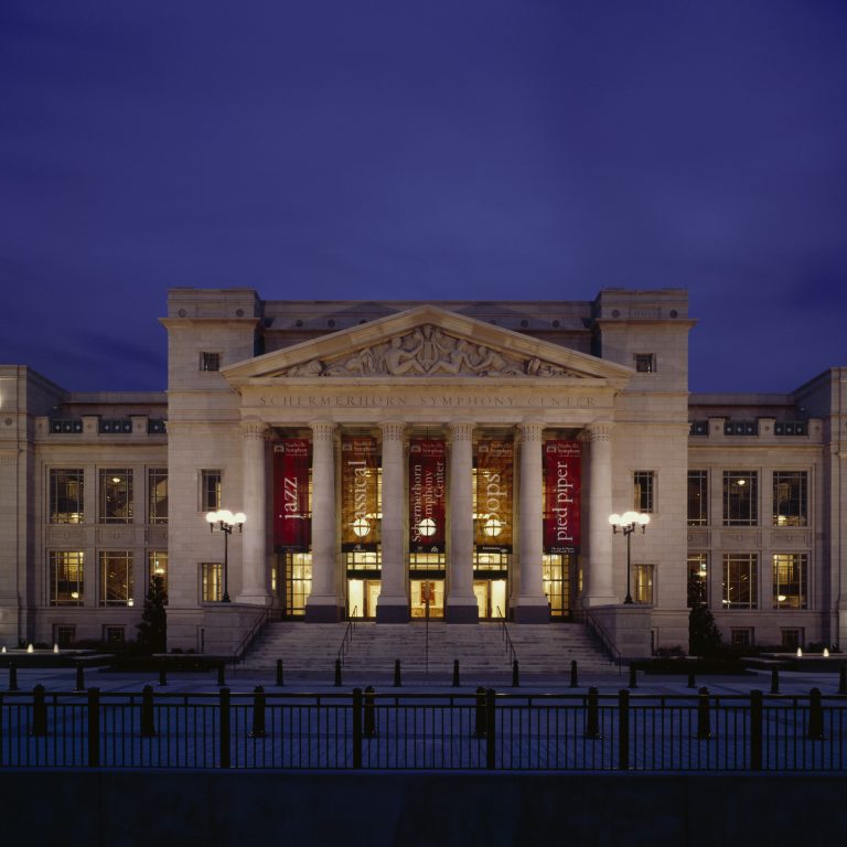 A large, illuminated neoclassical building with columns and banners at night, featuring Scherrmerhorn Symphony Center engraved above the entrance.
