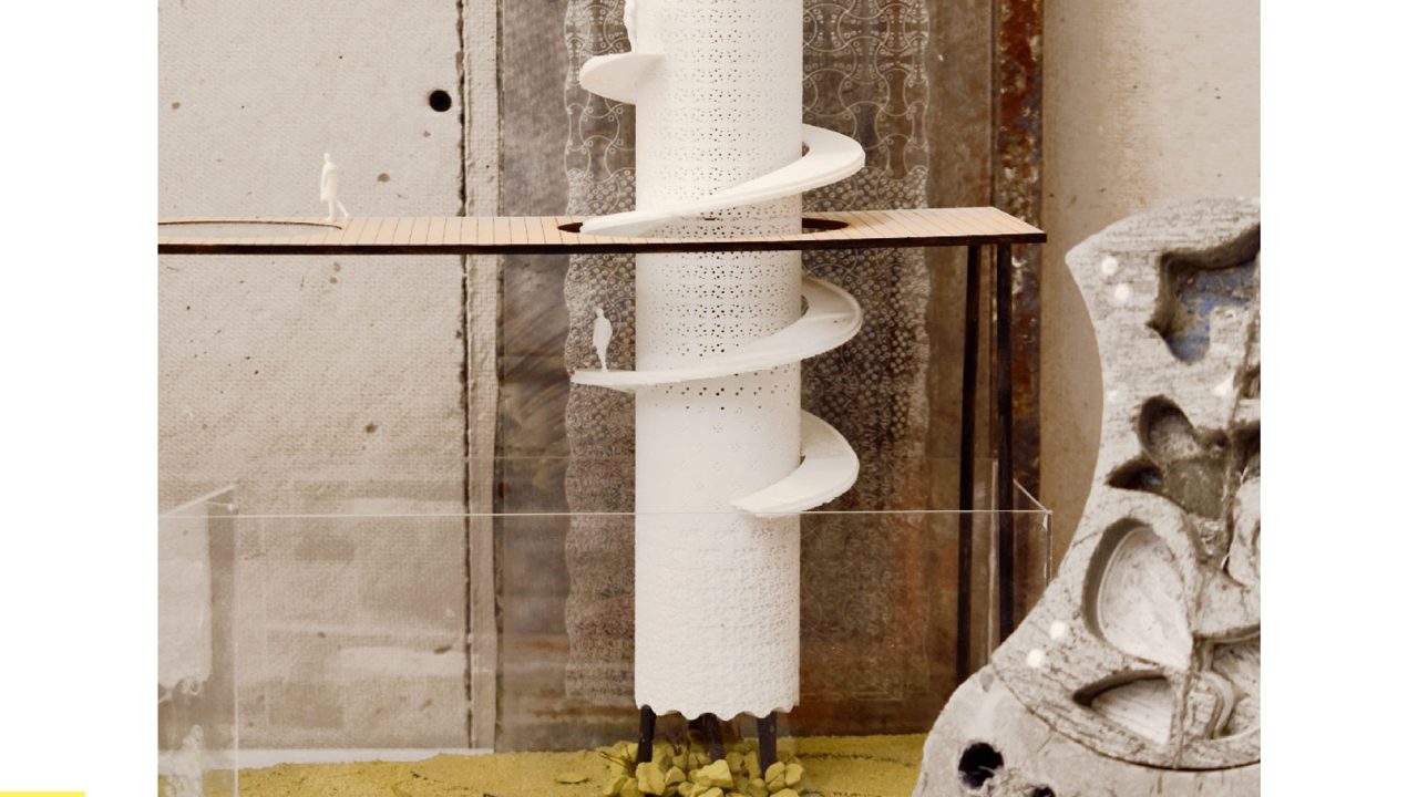 A cylindrical architectural model with perforated surfaces and a spiral ramp stands amid stone textures and construction materials, displayed on a sandy base.