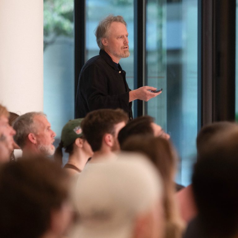 A man stands and speaks while holding a remote in front of a seated audience in a modern indoor setting.
