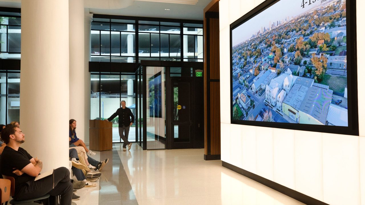Several people sit in a modern lobby watching a large screen displaying an aerial view of a cityscape with autumn trees.