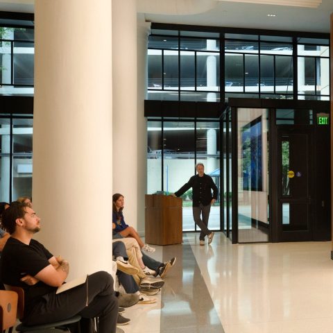 Several people sit in a modern lobby watching a large screen displaying an aerial view of a cityscape with autumn trees.
