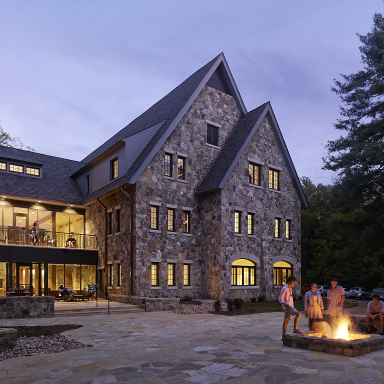 A large stone building with multiple windows and peaked roofs; several people are gathered around a fire pit in the courtyard at dusk.